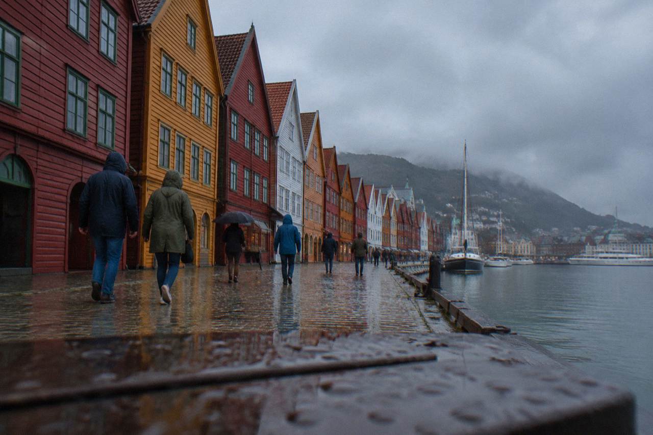 People in jeans and rain jackets walking along the colorful wooden wharf buildings of Bryggen in Bergen on a drizzly day