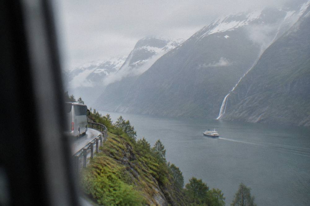 A coach on a narrow mountain road above a Norwegian fjord with a waterfall on the far shore