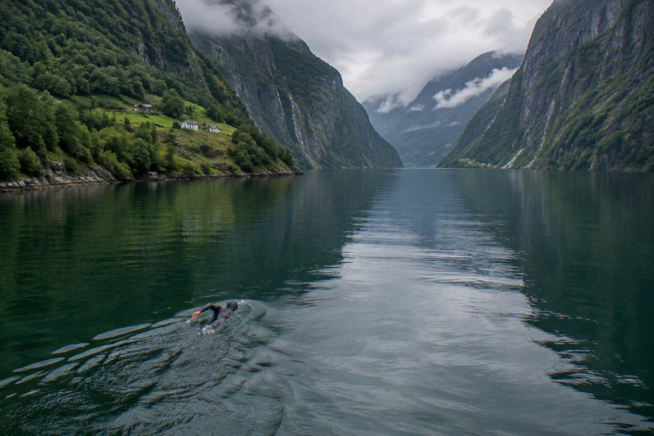 A swimmer in a wetsuit gliding through calm green fjord water beneath steep forested mountains in western Norway