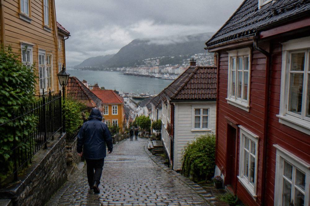 A person in a rain jacket walking up a wet, steep cobblestone lane between colourful wooden houses in Bergen, Norway.