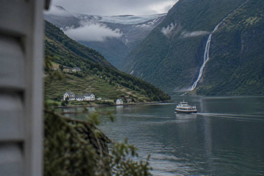 A waterfall dropping down a green cliff face toward a fjord with a tour boat on the water
