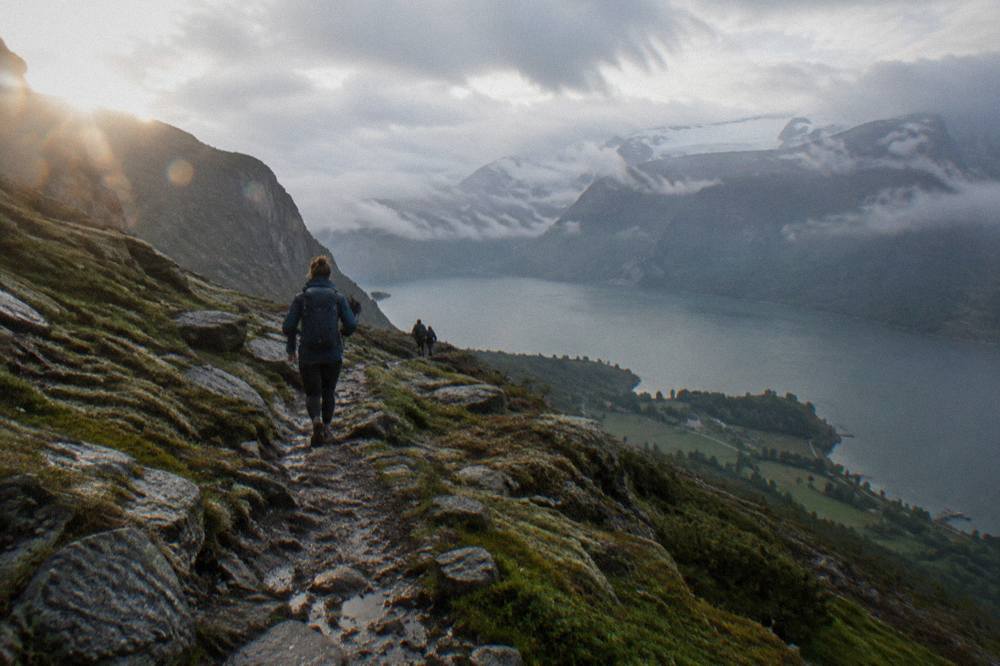 Hikers on a rocky mountain trail above a fjord, with a cliff ledge and dark water visible in the distance below clouds