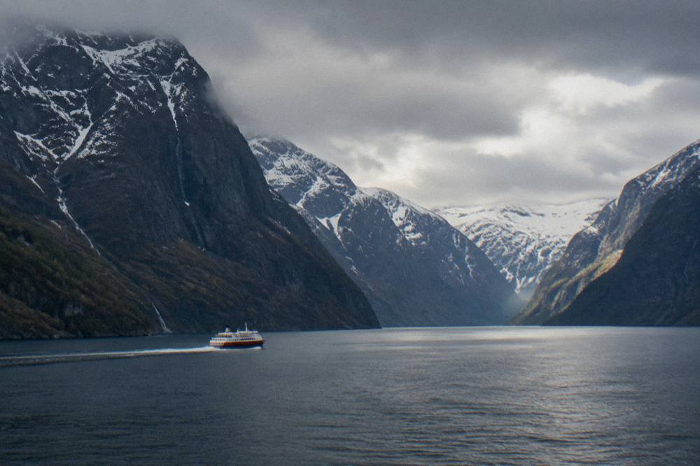 A red and white passenger boat travelling along a narrow fjord between snow-dusted cliffs