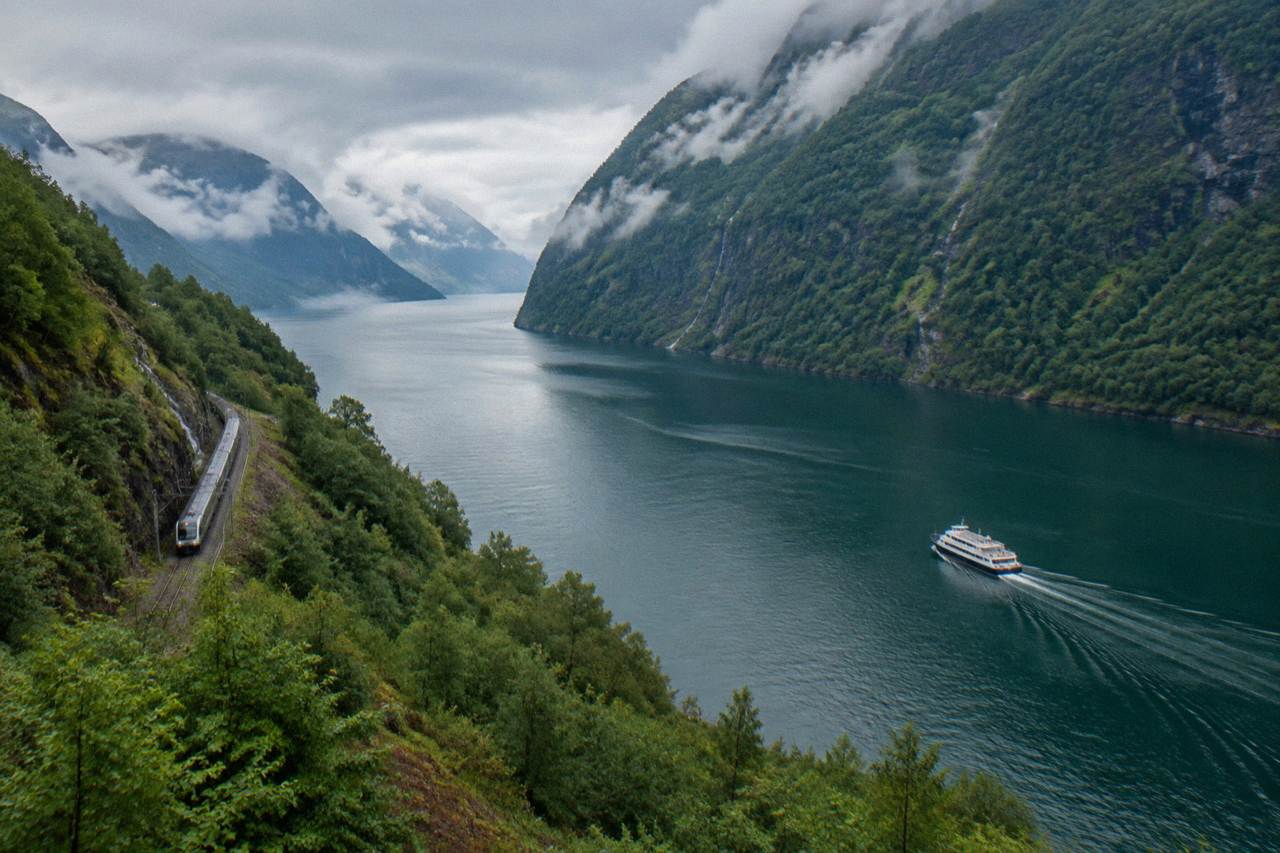 A scenic passenger boat travelling along a narrow Norwegian fjord with steep green mountainsides rising above the water