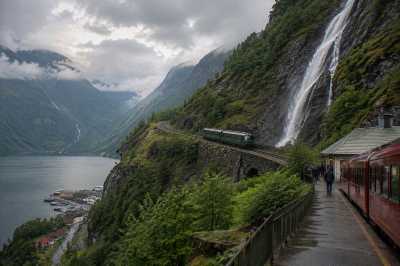 A red passenger train waiting at a small Norwegian mountain station beside a deep valley