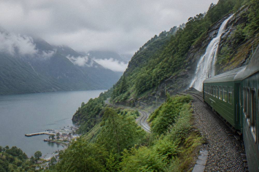 A steep railway track winding down a wooded mountainside toward a fjord village below