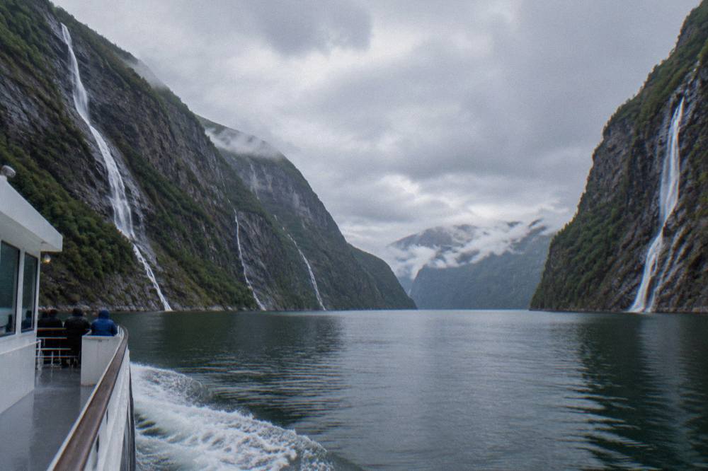 A tour boat travelling along a narrow fjord channel with steep grey rock cliffs on both sides and waterfalls running down them