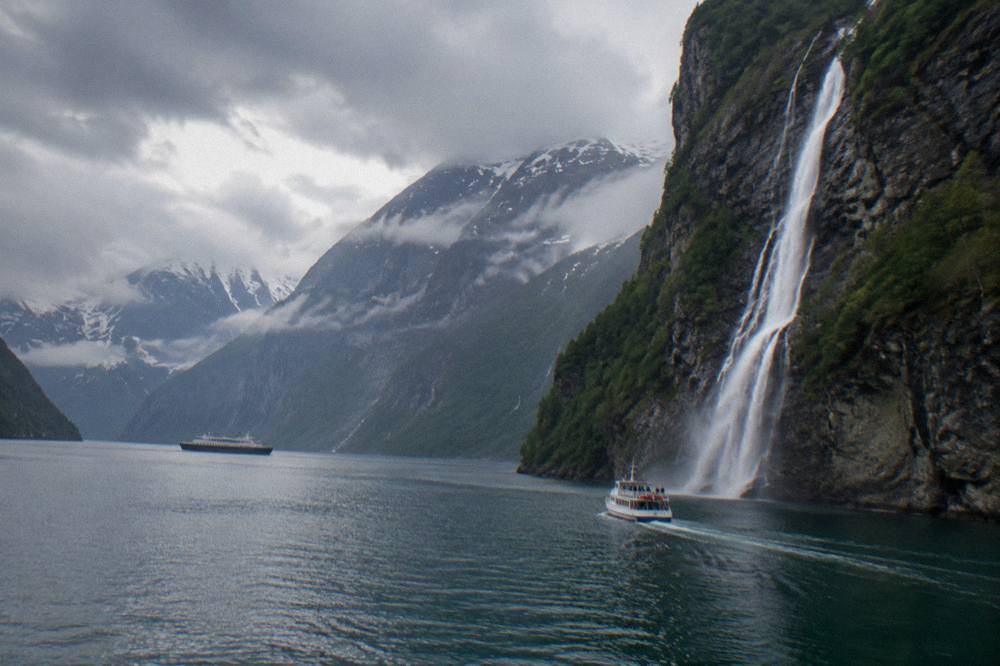 A small passenger boat cruising close to a waterfall dropping down a cliff face into a narrow Norwegian fjord