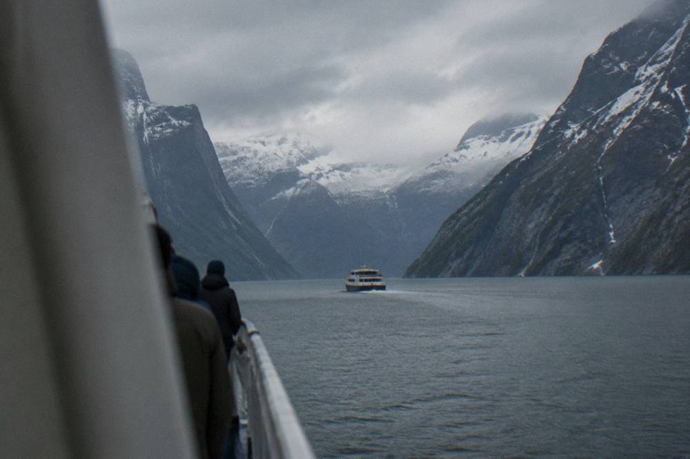 A small cruise boat on dark fjord water between steep cliffs, passengers on the forward deck