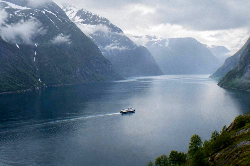 A small boat crossing calm fjord water beneath steep mountain walls under a partly cloudy sky