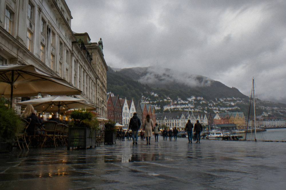 A wide stone square in central Bergen with classical facades, outdoor cafe seating and people walking between shop fronts