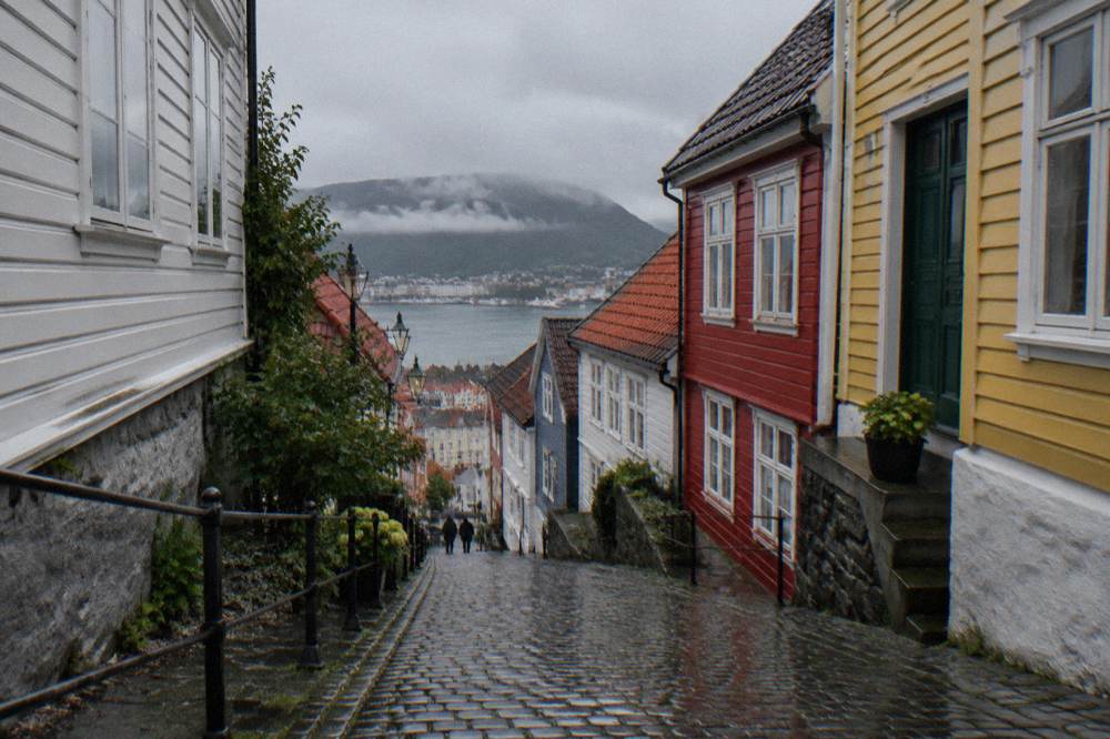 A narrow sloping lane in Bergen with colorful wooden houses on both sides and wet cobblestones reflecting the light