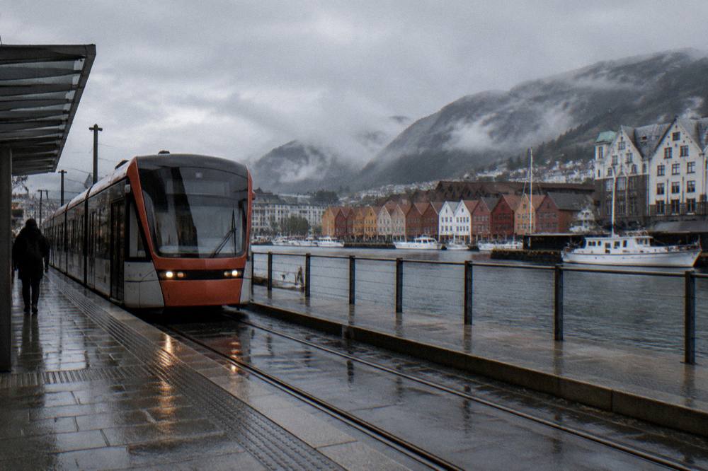 A modern light rail tram stopping at a central Bergen station platform with the fish market and harbor buildings in the background