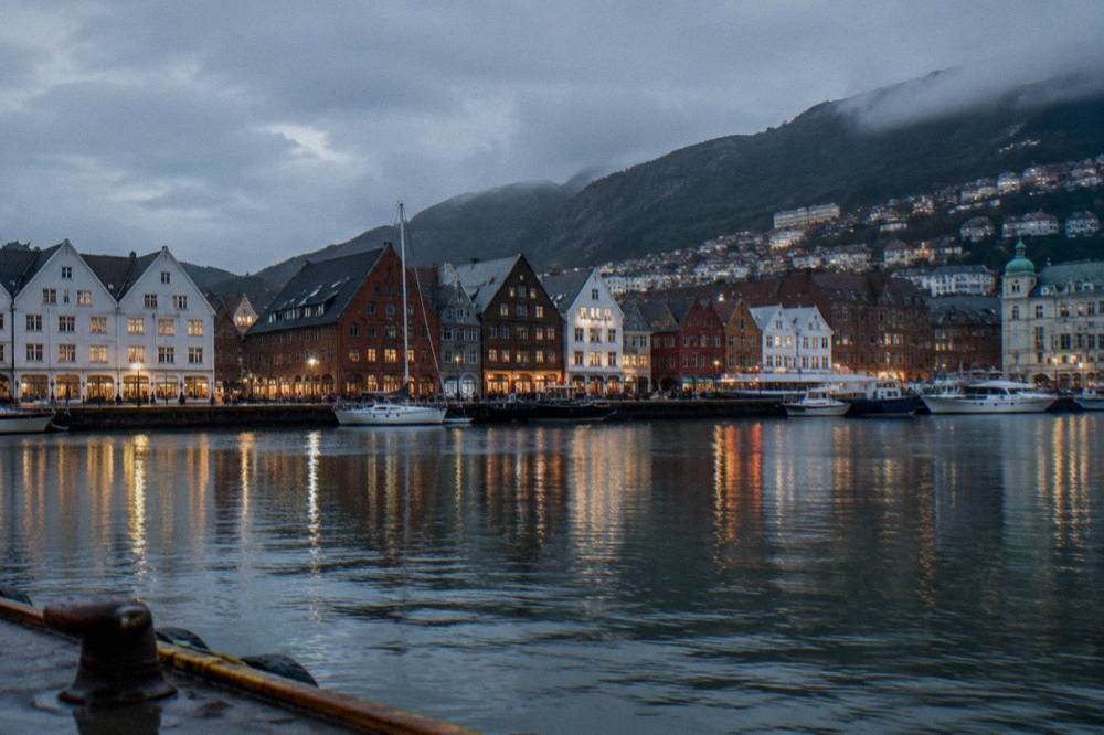 A harborside row of hotels and historic warehouses in Bergen seen across the water at dusk with lights reflecting in the harbor