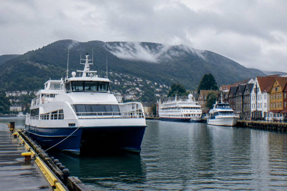A passenger ferry and small cruise boats moored at stone quays in central Bergen with forested mountains rising behind the harbor