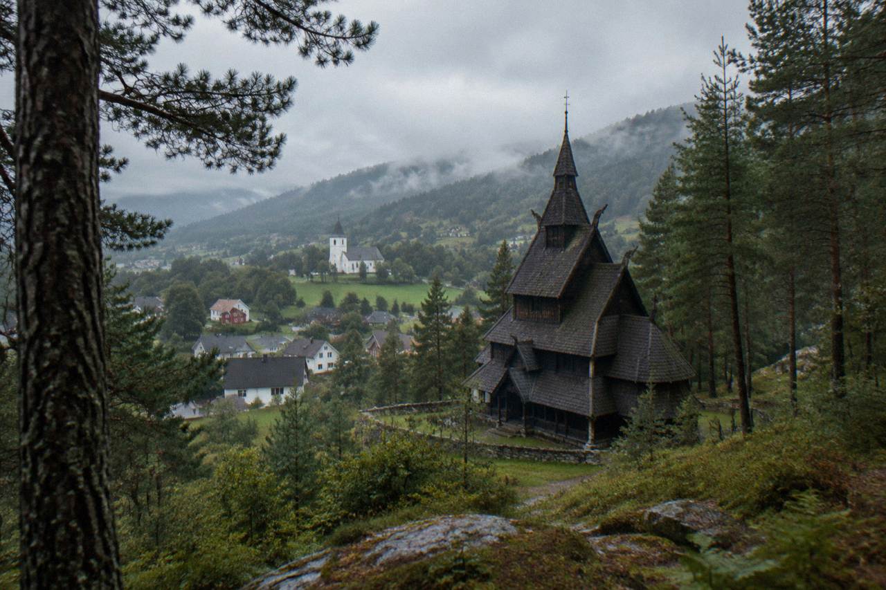 A wooden stave church with tiered roofs standing among tall pine trees on a quiet hillside