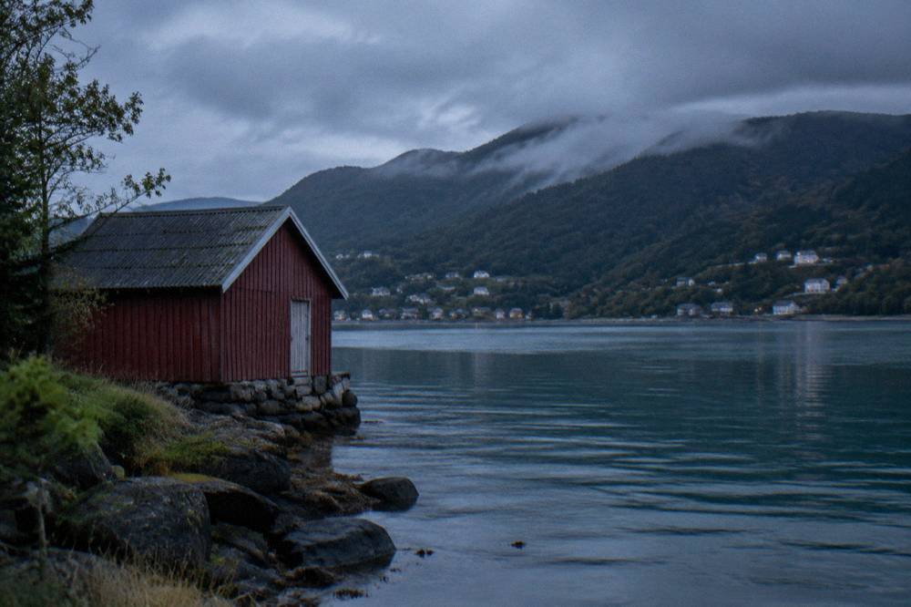 A lake shore with a small wooden boathouse and forested hills behind it