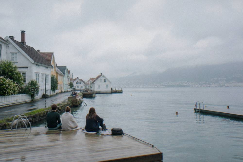 People sitting on a wooden pontoon by a harbour bath with the fjord behind