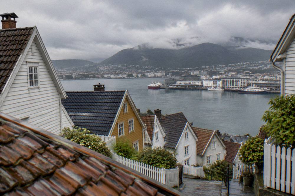 A viewpoint above wooden rooftops looking south across a harbour toward a wharf and mountains