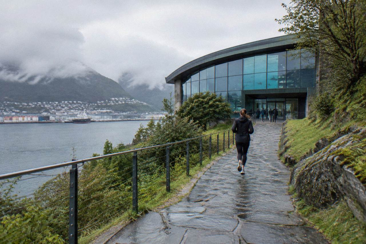 An aquarium building on a green hillside above the water in Bergen with a stone path leading to the entrance