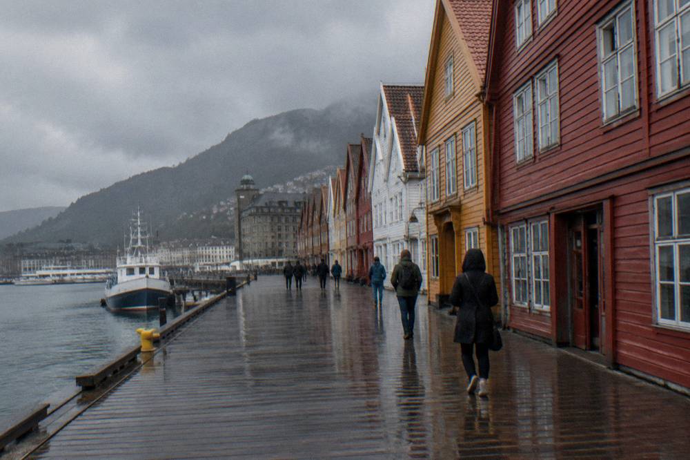 People walking past painted wooden trade buildings at a historic wharf in a Nordic harbour city