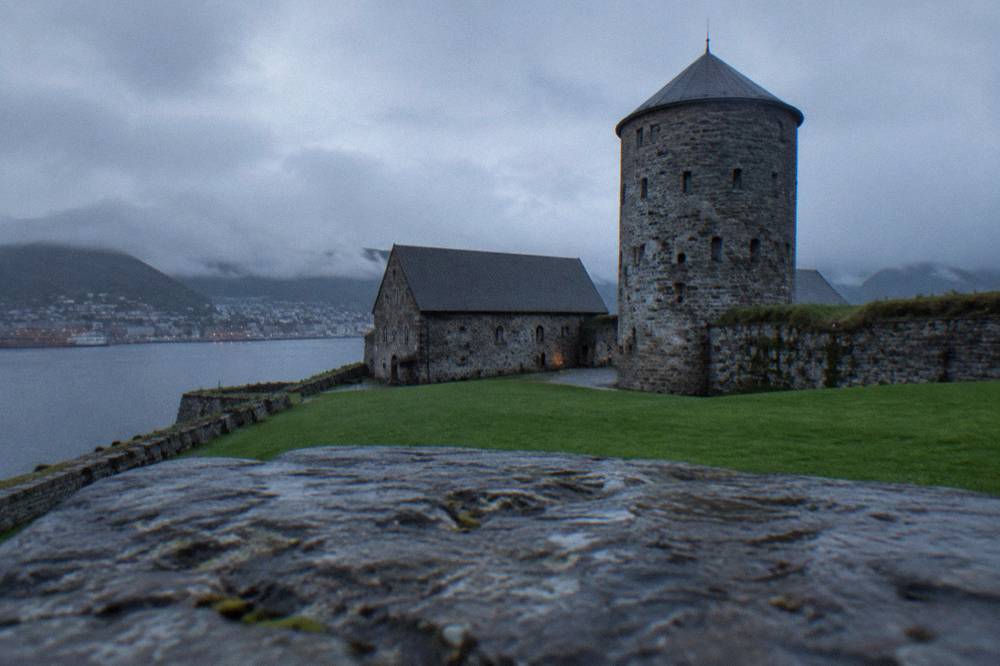 A stone tower with small window slits rising above a fortress wall with a green lawn in the foreground