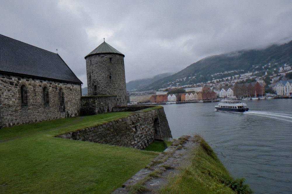 A sightseeing boat moving across a harbour toward colourful buildings and hills under a cloudy sky