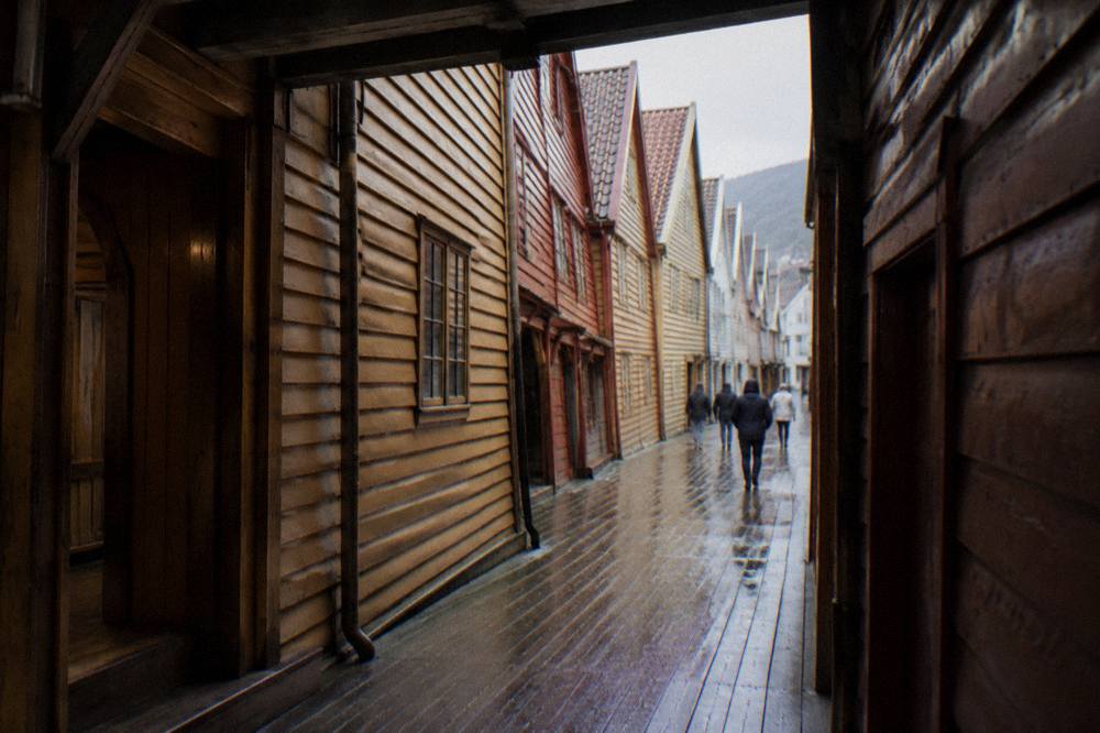 A narrow covered wooden lane between two warehouse buildings on Bryggen, with daylight falling between the tall timber walls.