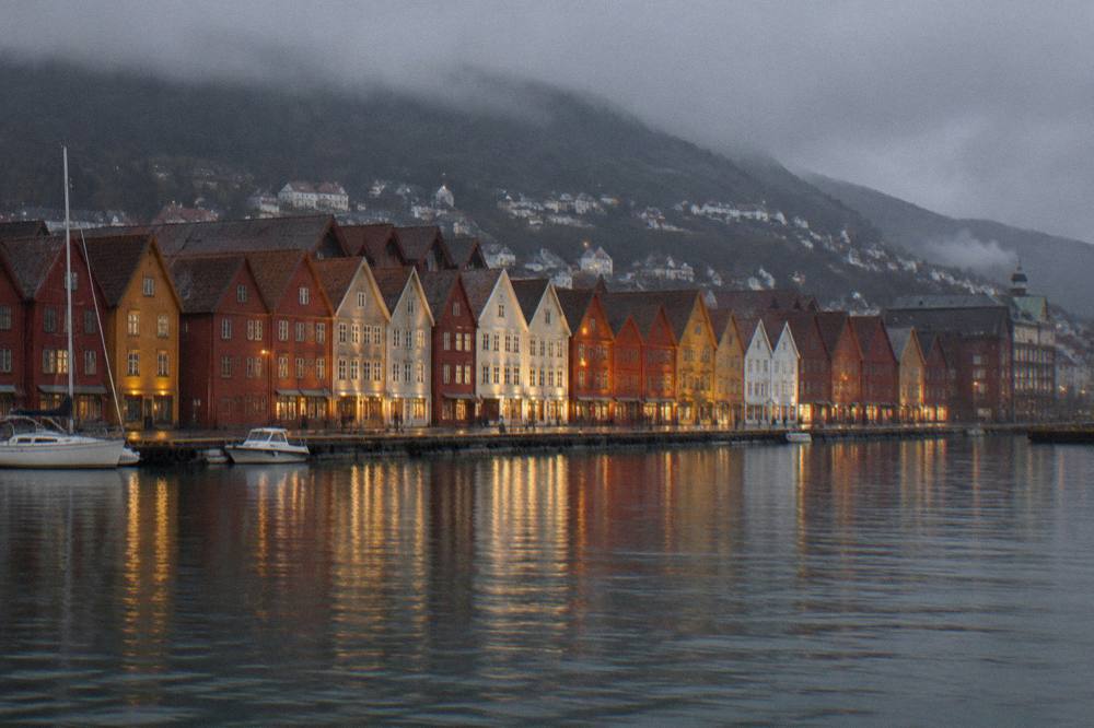 Bryggen seen from across the harbour at dusk, with the timber gables lit by warm lights against a dark sky.