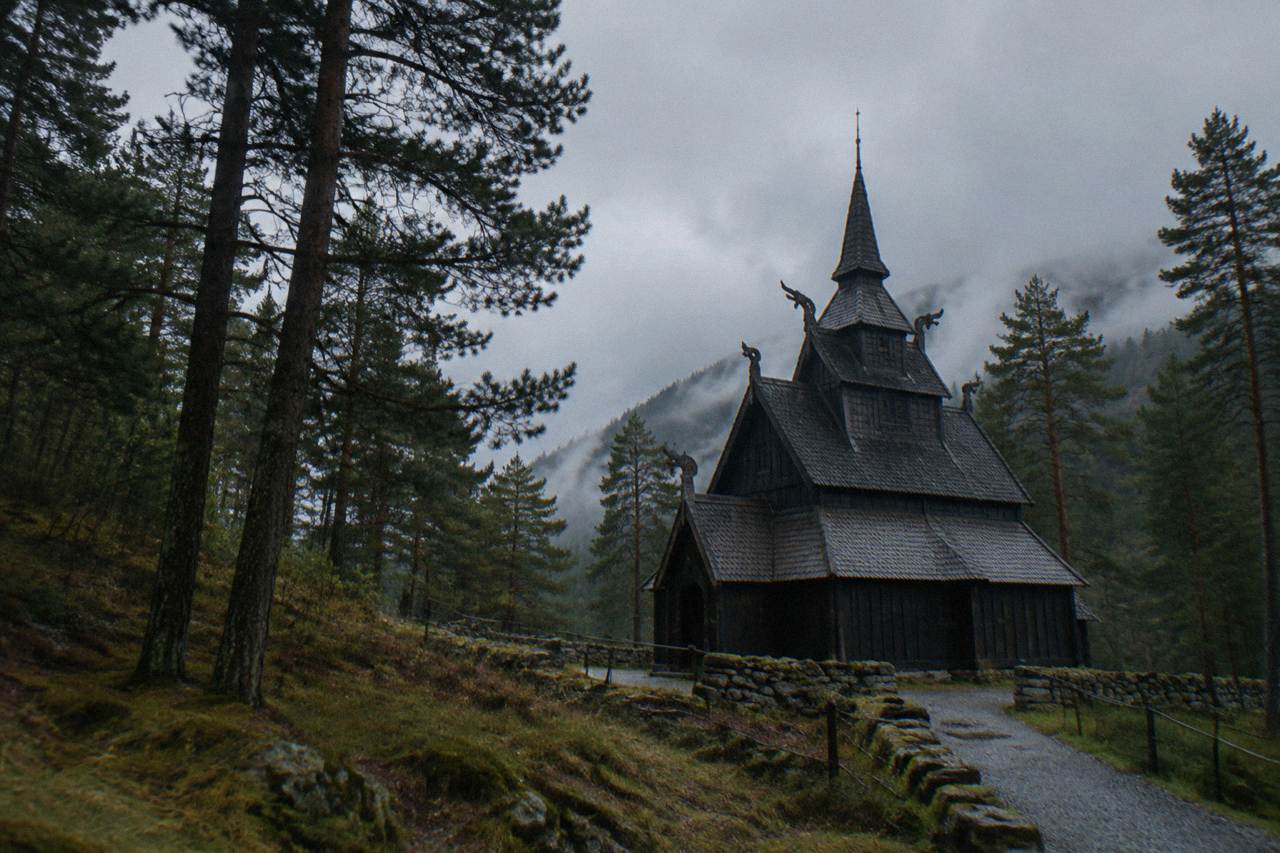 A dark wooden stave church with a tiered roof and carved gables standing among tall trees