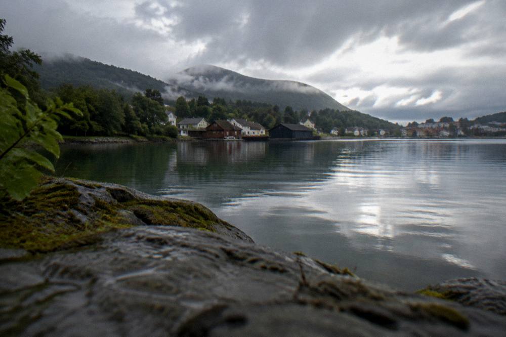 A calm lake surrounded by trees with wooden buildings on the far shore under a grey sky
