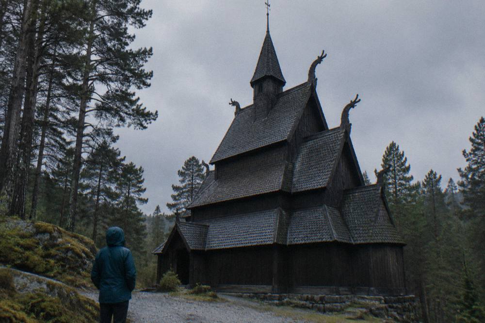 A visitor in a rain jacket standing on a gravel path looking up at the tiered wooden roof of the stave church
