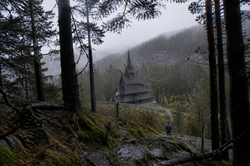 The stave church seen at a distance through a frame of tall dark trees with soft evening light