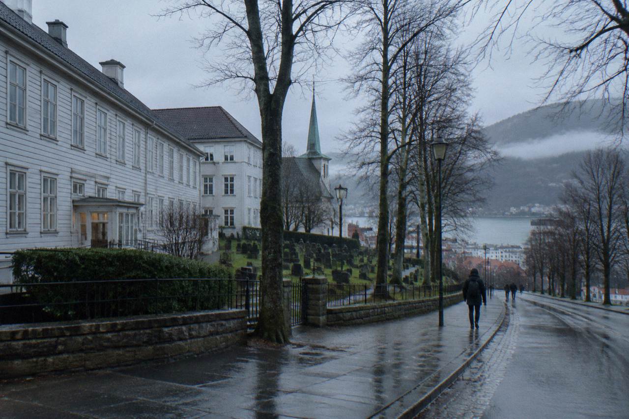 Wooden hospital buildings and a quiet churchyard beside a tree-lined street in central Bergen