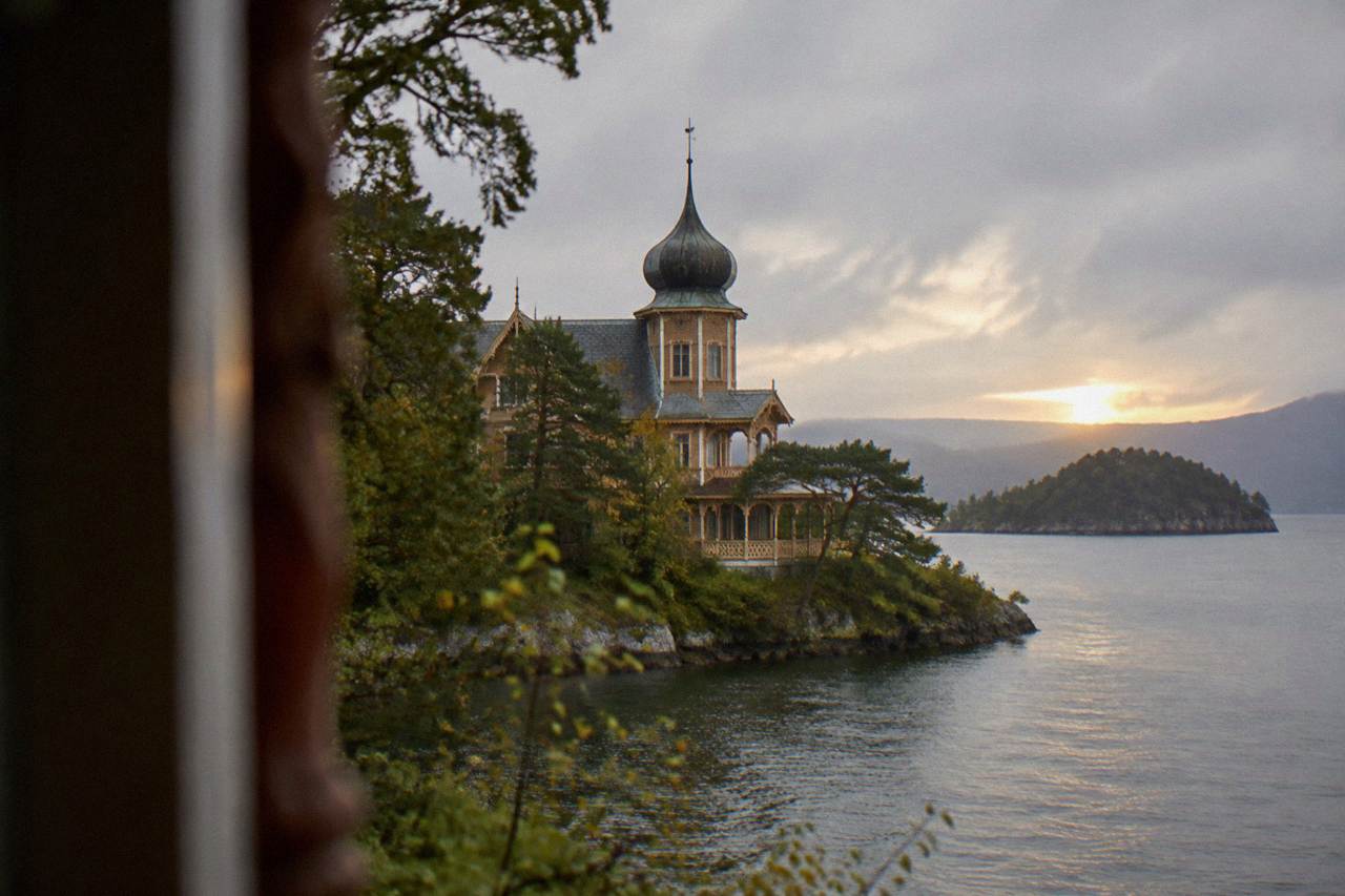 A wooden villa with ornate towers and verandas standing among trees on a small Norwegian island, with calm water in the background.