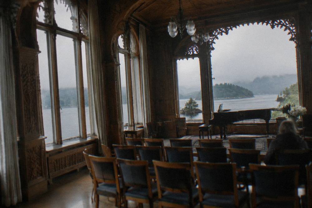 An ornately carved wooden music room with tall windows, a piano and rows of chairs facing a small performance space.