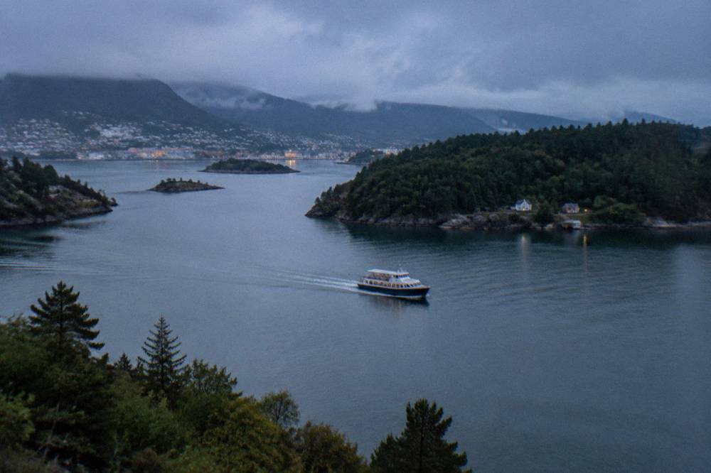 A small passenger boat crossing calm grey-green water between forested Norwegian islands near Bergen.