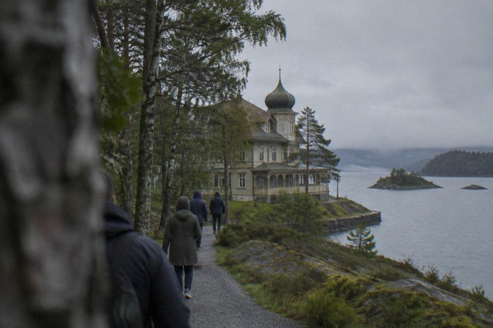 Visitors walking a gravel path through birch and pine woodland toward a wooden villa on a Norwegian island shoreline.