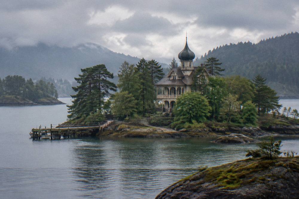 A quiet inlet with a wooden jetty, low rocky shoreline and forested islands under a partly cloudy Norwegian sky.