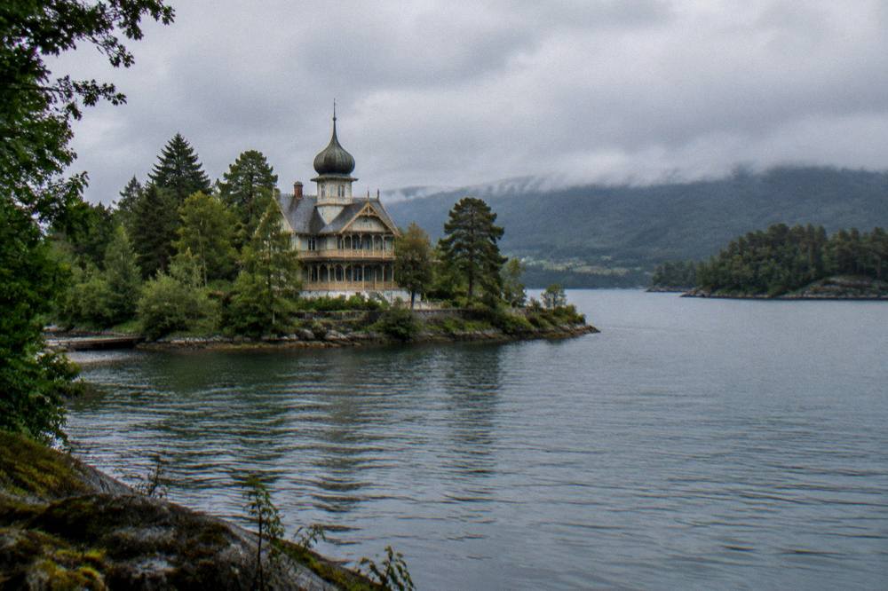 A panoramic view from a Norwegian island shoreline across dark water toward low forested hills at dusk.