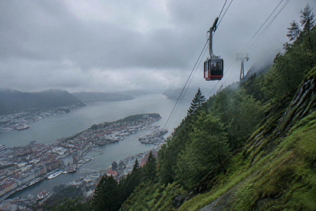 A cable car cabin travelling up a steep green mountainside above a fjord and city rooftops