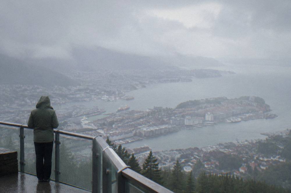 A person in a rain jacket standing on a summit platform overlooking a coastal city and fjord