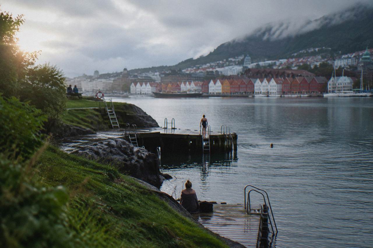 Wooden piers and ladders at a sea bath on a green peninsula in Bergen, with calm harbour water and coloured houses across the bay