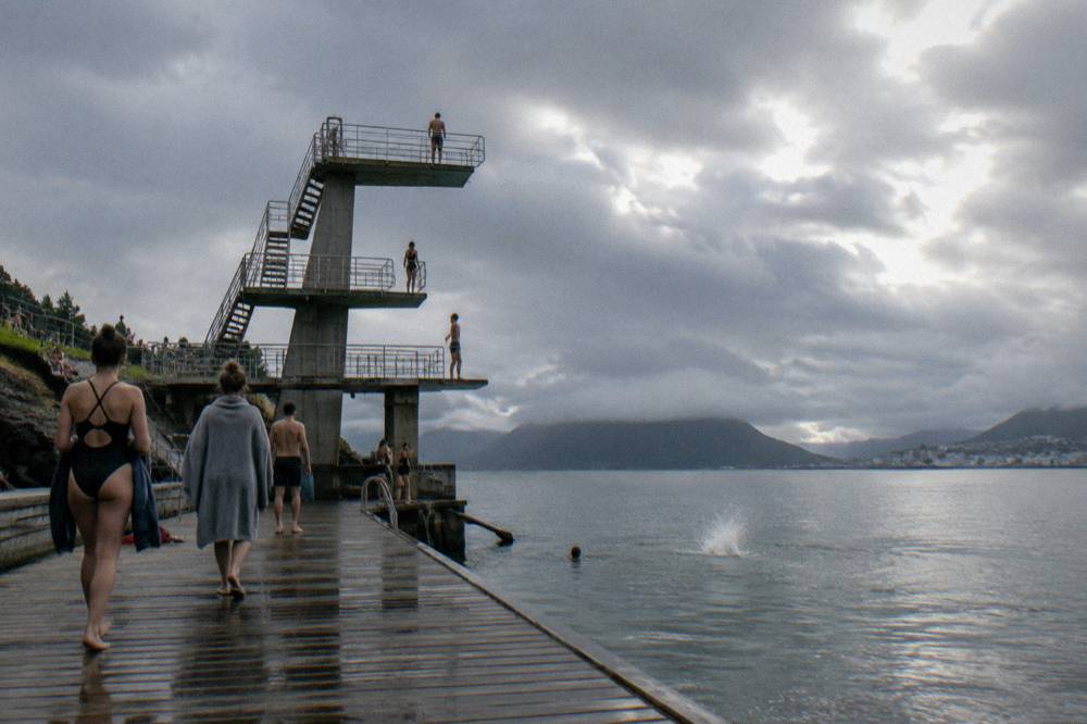 People in swimsuits and robes on a wooden pier at a Norwegian sea bath, grey sky and calm fjord water beyond