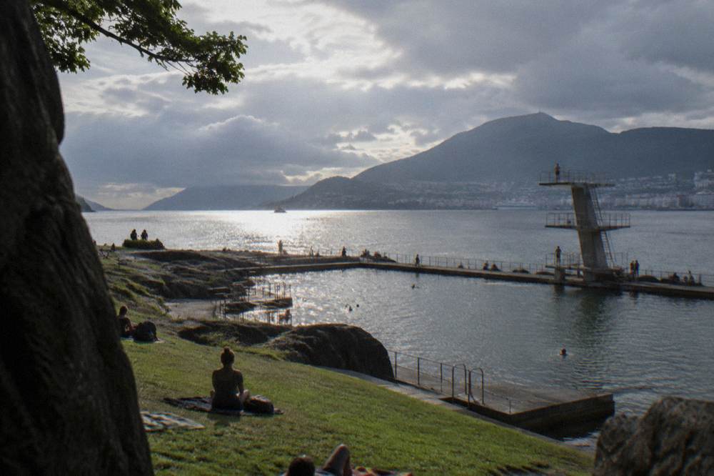 A calm summer evening over a Norwegian harbour peninsula, low sun on the water and mountains behind the city