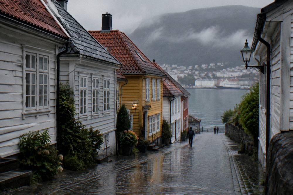 A quiet cobbled lane of low wooden houses on a peninsula in Bergen, wet pavement reflecting the facades