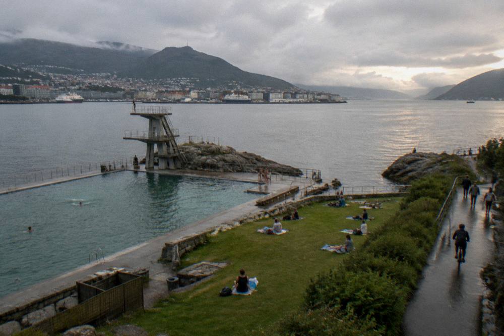 A cyclist and pedestrians on a waterfront path beside a fjord, green hills rising behind a Norwegian city