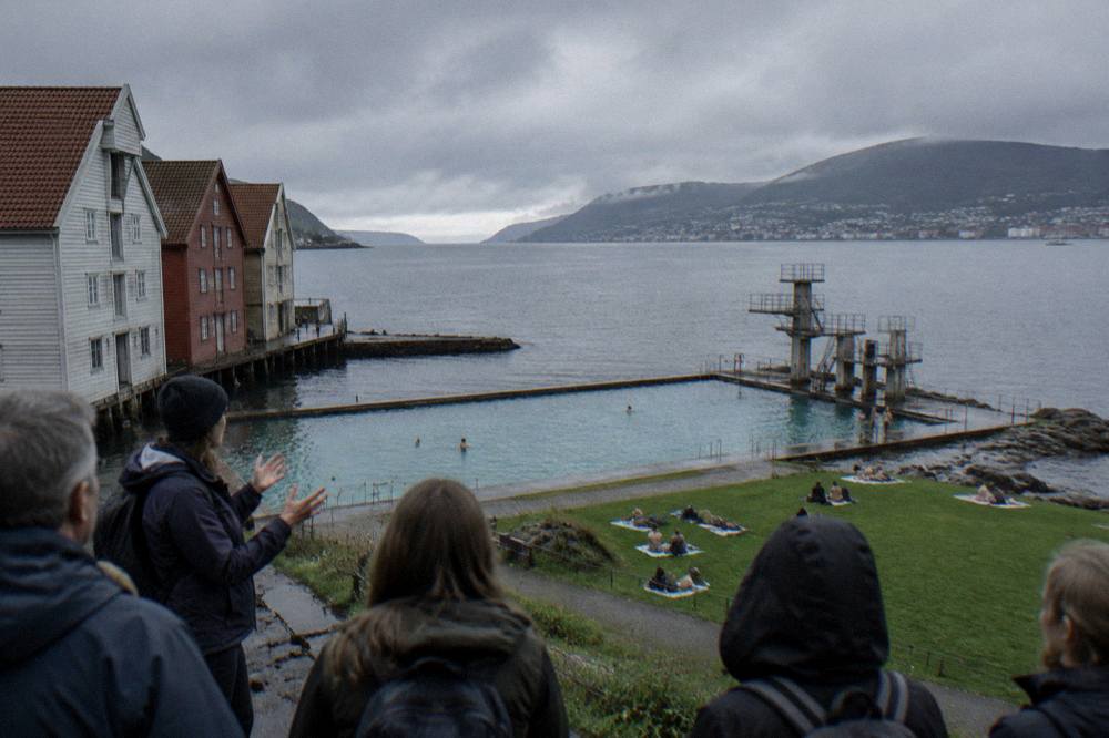 A guide speaking to visitors beside old wooden warehouses at the water's edge in a Norwegian harbour city