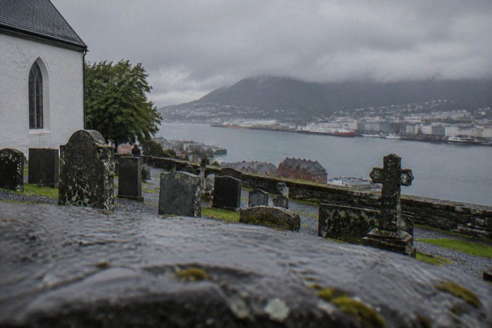 Gravel churchyard with old headstones and benches, viewed against Bergen harbor water