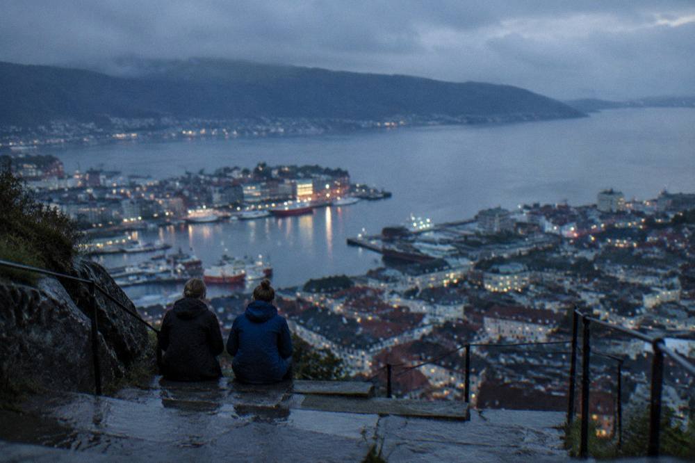 Two walkers resting at the top of a stone stairway above a city with fjord water beyond